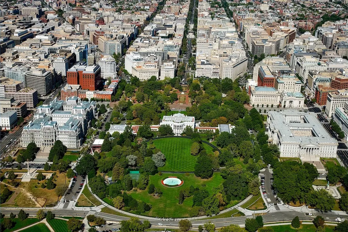 Aerial view of the White House surrounded by trees and government buildings in Washington, D.C., with city streets and densely packed buildings extending into the distance.
